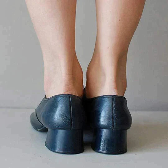 Chaussures en cuir noir à talons bas portées par une personne debout sur un sol gris. Mode élégante, confort, style classique.