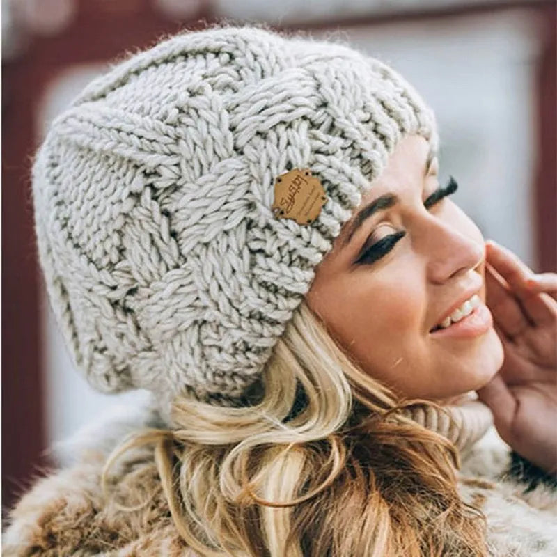 Femme souriante portant un bonnet en laine tricoté gris avec motif torsadé, étiquette en cuir, cheveux blonds bouclés, mode hiver élégante.