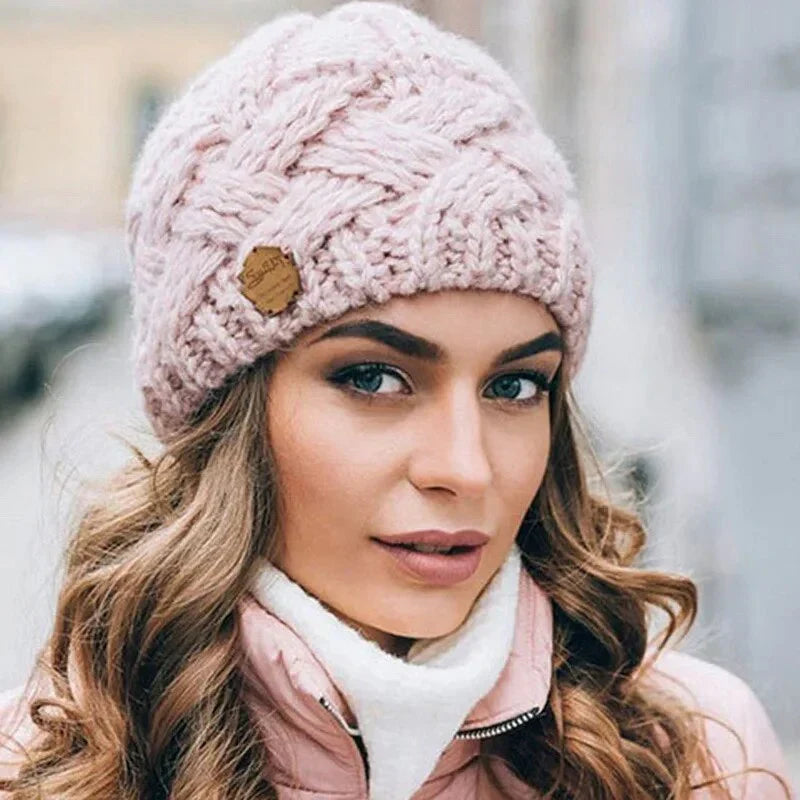 Femme portant un bonnet en laine rose tricoté, avec un motif torsadé, étiquette en cuir, manteau rose, cheveux bouclés, mode hiver élégante.