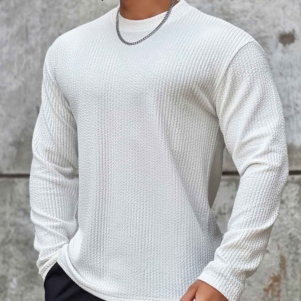 Homme portant un pull blanc texturé à manches longues, chaîne argentée, fond béton. Mode masculine, style décontracté, tendance automne-hiver.