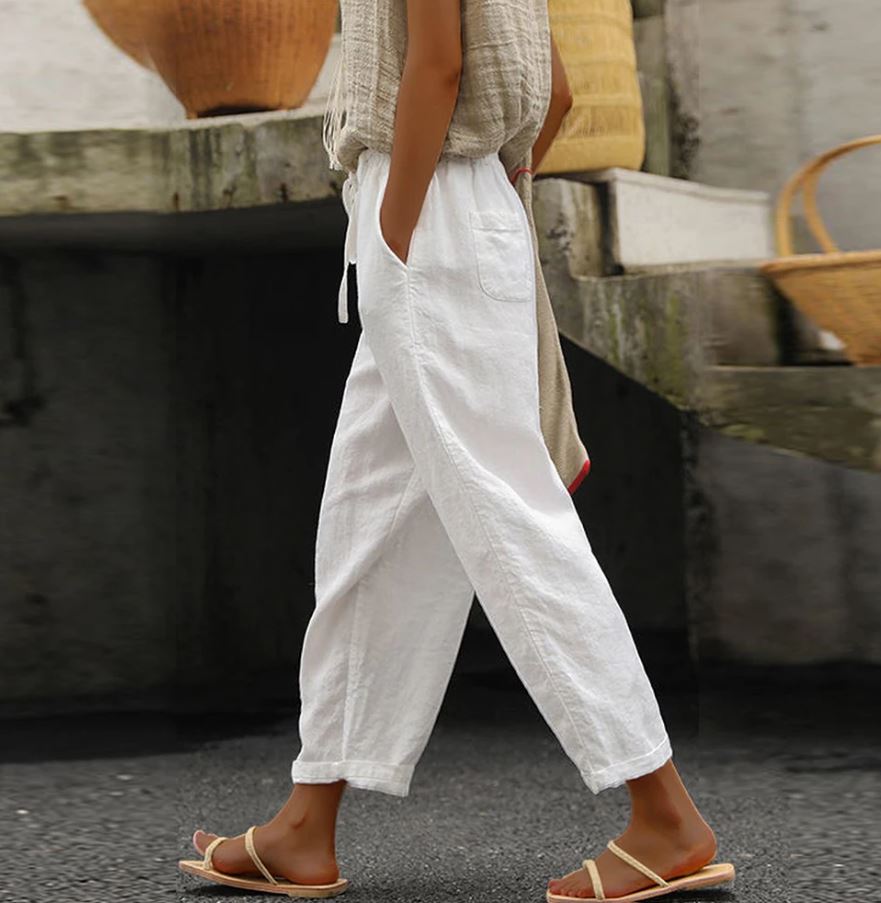 Femme portant un pantalon blanc en lin, sandales beiges, marchant sur un trottoir. Mode estivale décontractée, style minimaliste.