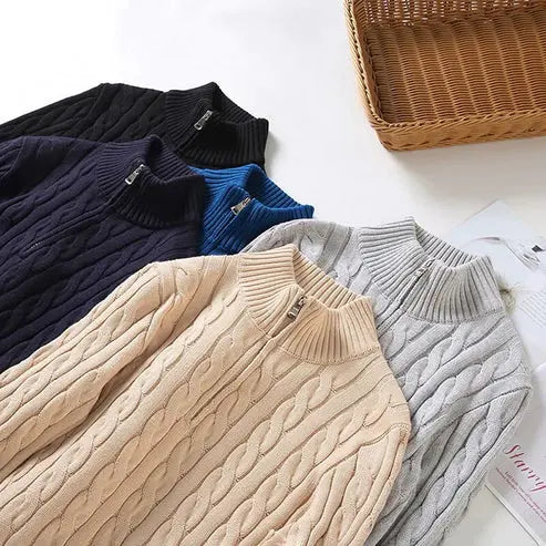 Cinq pulls en maille torsadée de différentes couleurs (noir, bleu, beige, gris) posés à côté d'un panier en osier, mode automne-hiver.