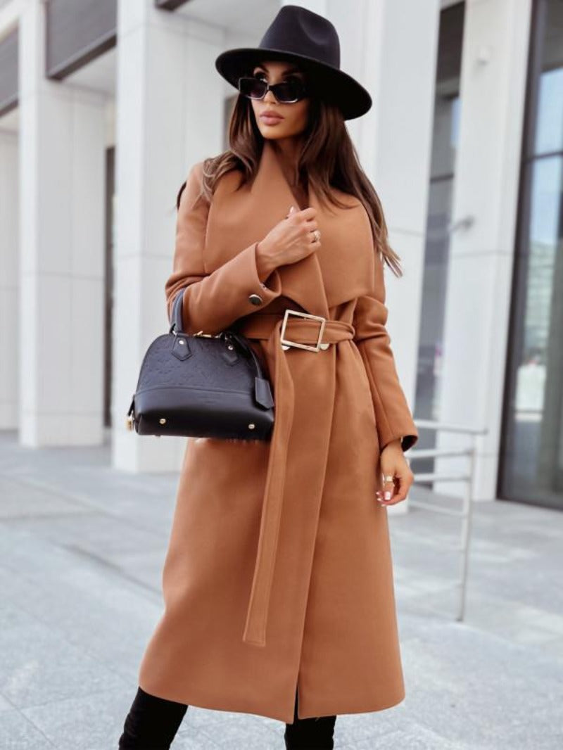Femme élégante en manteau camel ceinturé, chapeau noir et lunettes de soleil, tenant un sac à main noir devant un bâtiment moderne.