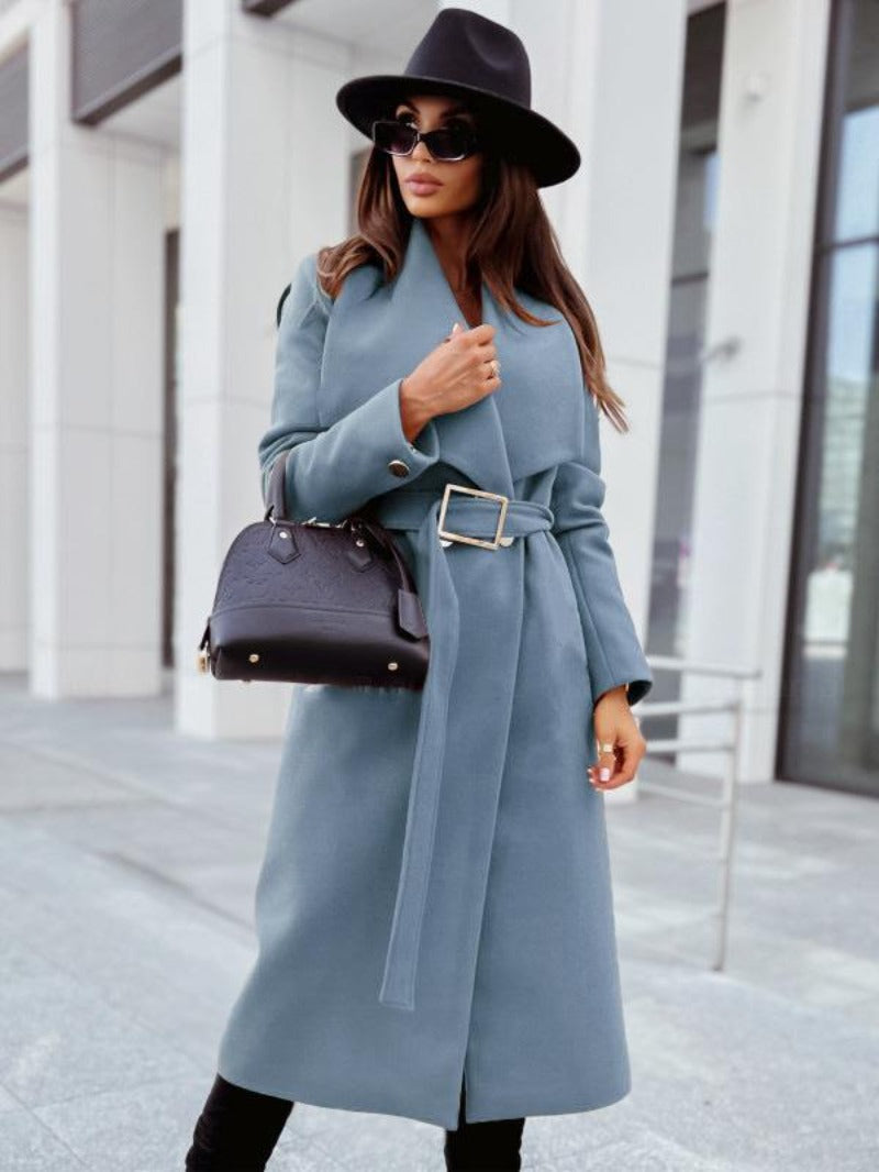 Femme élégante en manteau bleu ceinturé, chapeau noir et lunettes de soleil, tenant un sac à main noir devant un bâtiment moderne.