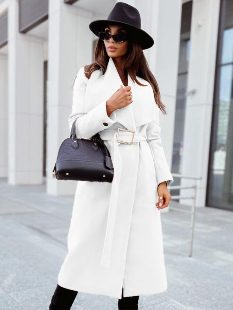Femme élégante en manteau blanc, chapeau noir et lunettes de soleil, tenant un sac à main noir devant un bâtiment moderne. Mode chic et urbaine.