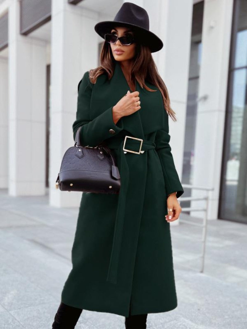Femme élégante en manteau vert foncé avec ceinture, chapeau noir et lunettes de soleil, tenant un sac à main noir devant un bâtiment moderne.