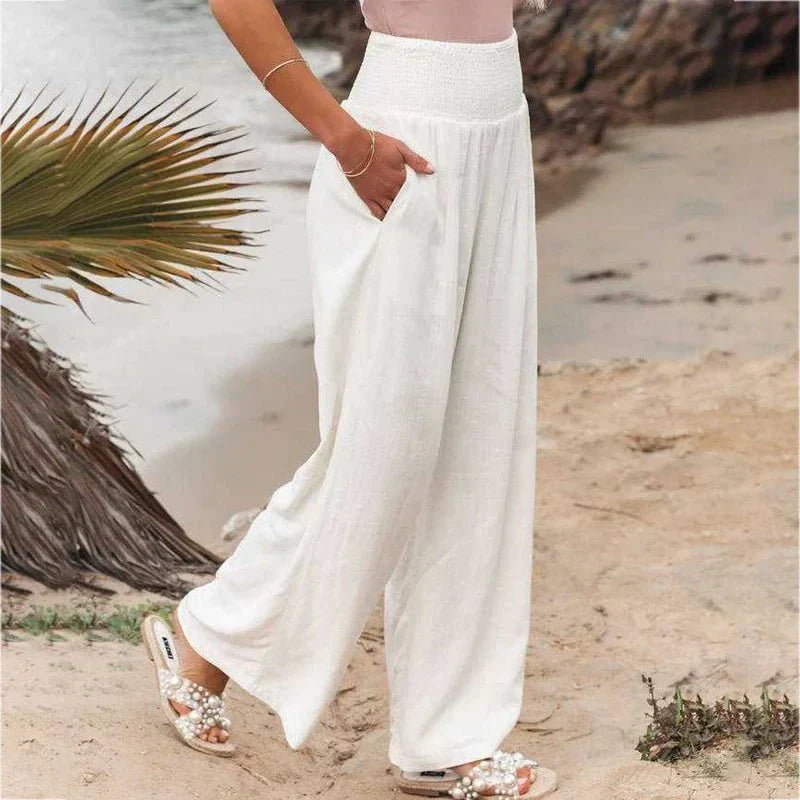 Femme portant un pantalon blanc ample en lin, debout sur une plage. Mode estivale, tenue décontractée, sandales perlées, style bord de mer.