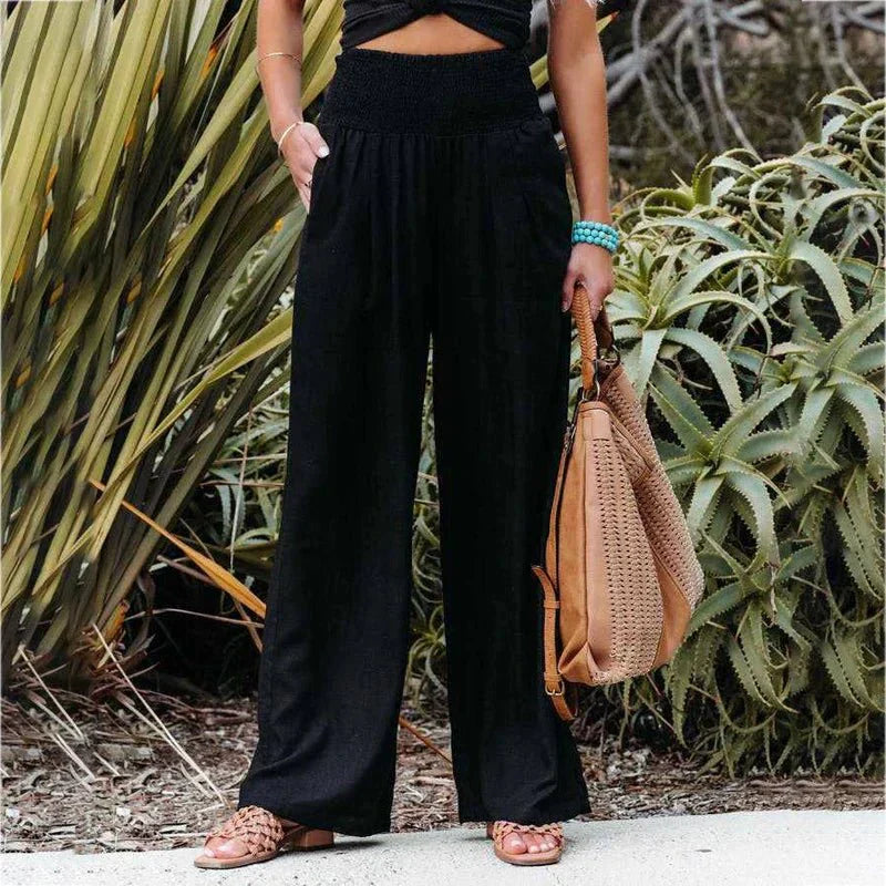 Femme portant un pantalon noir ample taille haute, sandales marron, et sac à main beige, posant devant des plantes vertes. Mode décontractée chic.