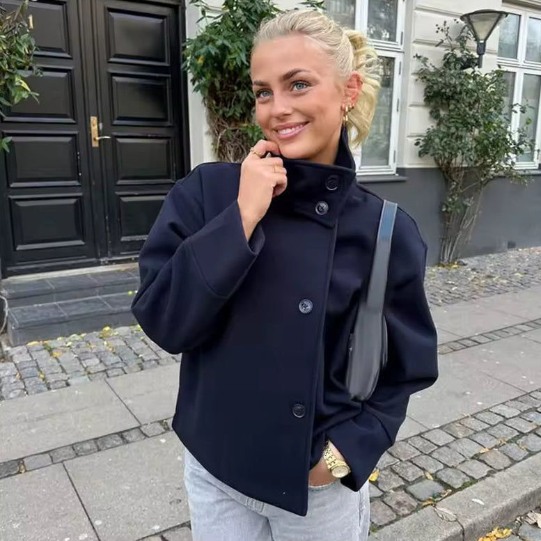 Jeune femme souriante en manteau noir élégant, posant dans une rue pavée devant une porte noire, cheveux blonds attachés, sac à l'épaule.