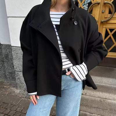 Femme en manteau noir et jean bleu devant une porte en bois, portant un pull rayé blanc et noir. Mode automne, style décontracté urbain.