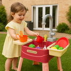 Petite fille jouant avec une cuisine en plastique rouge dans le jardin, versant de l'eau dans des tasses colorées. Jouet éducatif extérieur pour enfants.