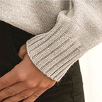 Manche de pull gris en tricot côtelé, main posée sur un pantalon noir. Mode automne-hiver, vêtement chaud et confortable.