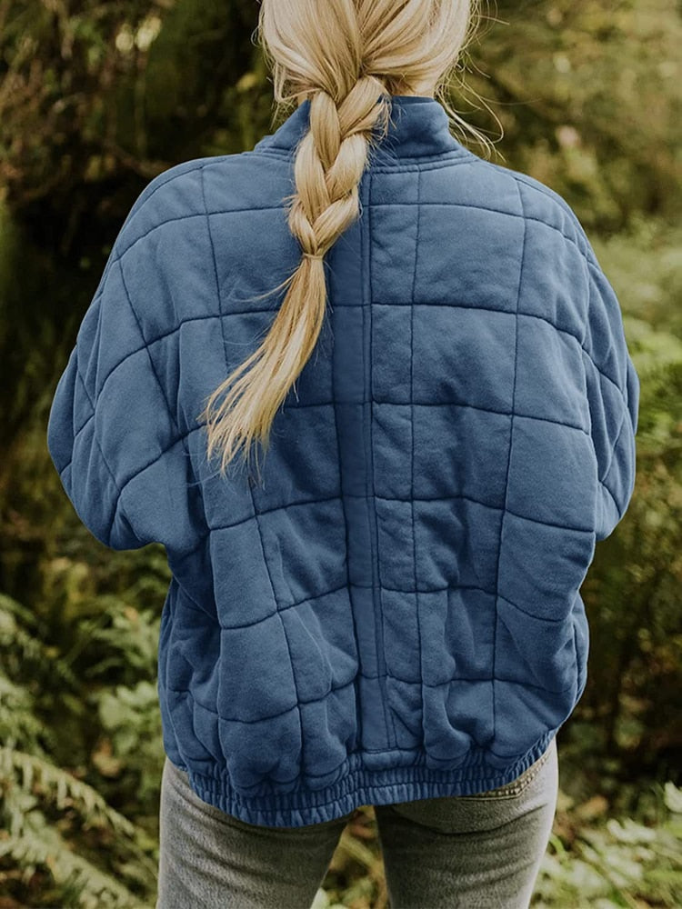 Femme de dos portant une veste matelassée bleue en forêt, cheveux blonds tressés. Mode automne, randonnée, confort, nature.