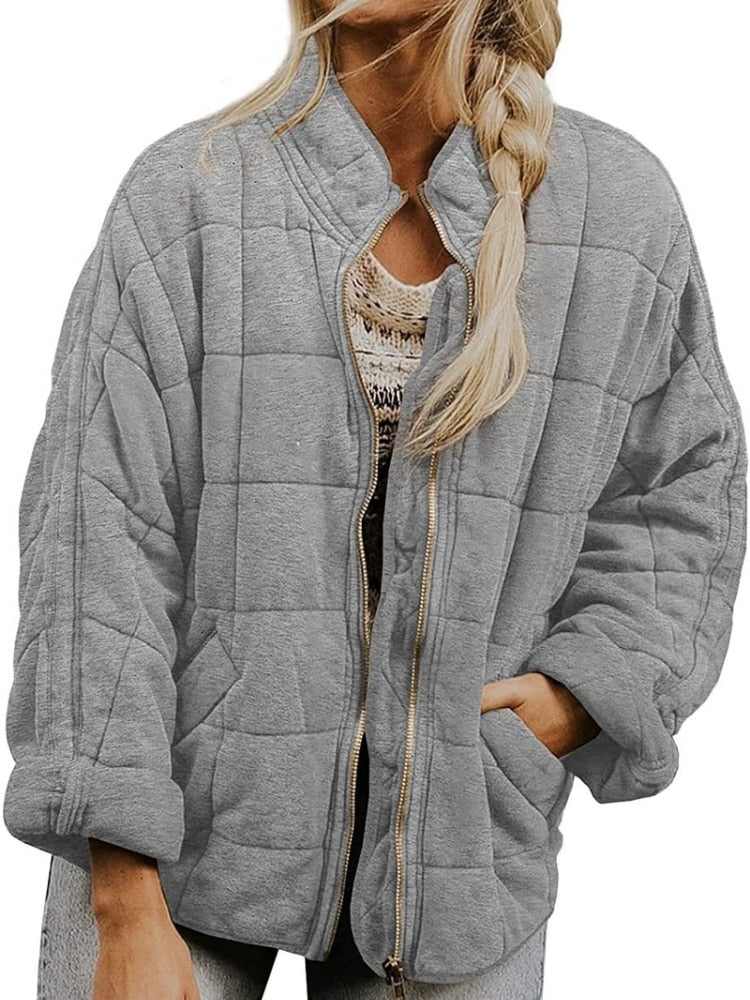 Femme portant une veste matelassée grise, manches retroussées, avec fermeture éclair dorée. Mode automne-hiver, style décontracté et tendance.