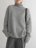 Femme portant un pull gris en laine à col roulé, manches longues, avec un sac noir. Mode automne-hiver, tenue élégante et confortable.
