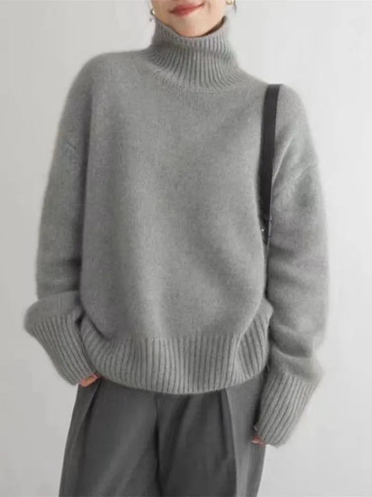 Femme portant un pull gris à col roulé en laine, manches longues, avec un sac noir. Mode automne-hiver, tenue décontractée élégante.