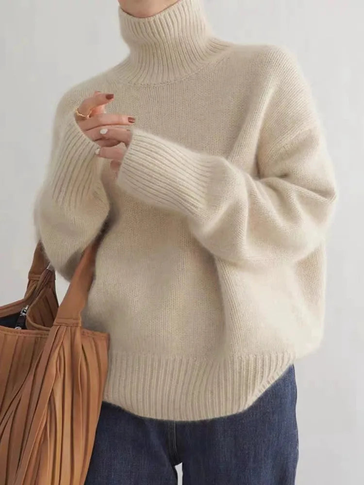 Femme portant un pull en laine beige à col roulé, manches longues, tenant un sac en cuir marron. Mode automne-hiver, style décontracté et élégant.