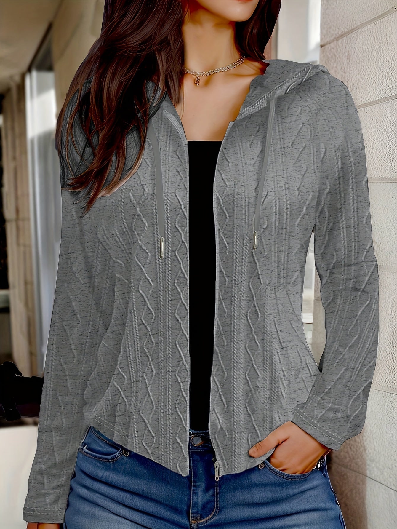 Femme portant un cardigan gris à capuche avec motif torsadé, manches longues, sur un haut noir, posant dans un couloir moderne. Mode automne-hiver.