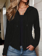 Femme portant un gilet noir à capuche en tricot torsadé, manches longues, avec un débardeur noir et un jean décontracté. Mode automne chic.