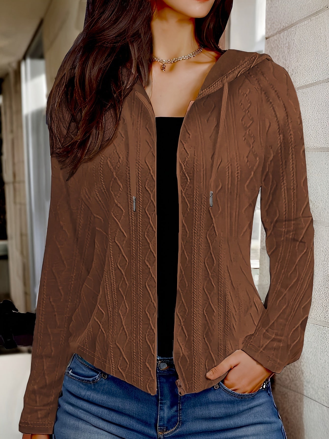 Femme portant un cardigan marron à capuche avec motif torsadé, sur un top noir et un jean bleu. Mode automne, style décontracté, tendance féminine.