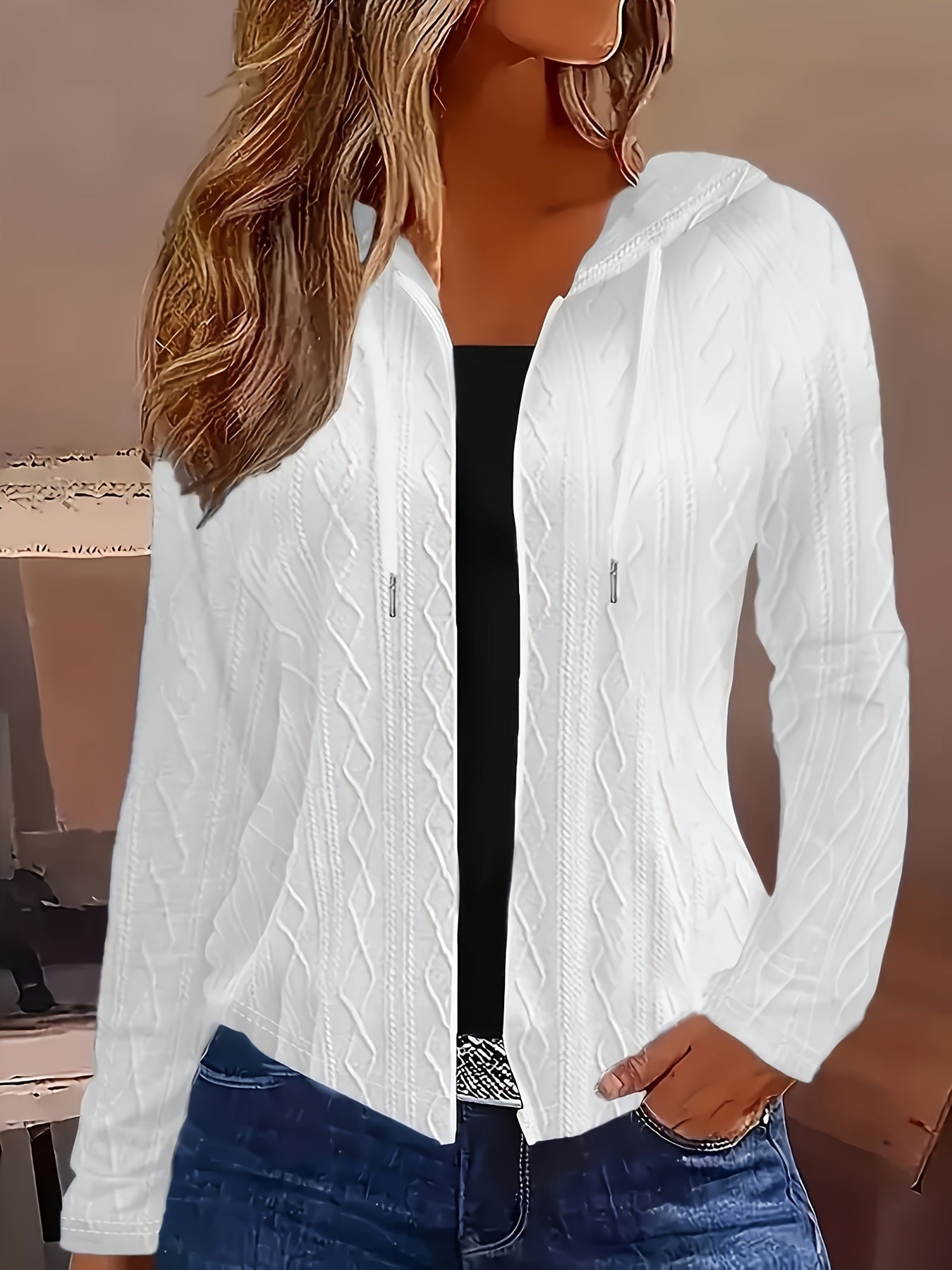 Femme portant un cardigan blanc à capuche en tricot torsadé, avec un haut noir et un jean bleu. Mode automne, style décontracté, tendance féminine.