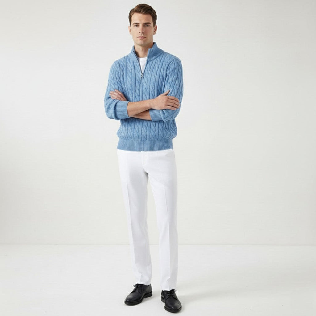 Homme portant un pull bleu en laine torsadée et pantalon blanc, posant debout sur fond blanc. Mode masculine élégante, tenue automne-hiver.