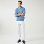 Homme portant un pull bleu en laine torsadée et pantalon blanc, posant debout sur fond blanc. Mode masculine élégante, tenue automne-hiver.
