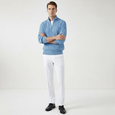Homme portant un pull bleu en laine torsadée et pantalon blanc, posant debout sur fond blanc. Mode masculine élégante, tenue automne-hiver.