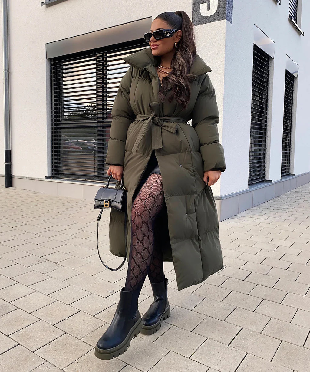 Femme portant un long manteau matelassé vert, collants à motifs, bottes noires épaisses et lunettes de soleil, tenant un sac à main noir devant un bâtiment moderne.