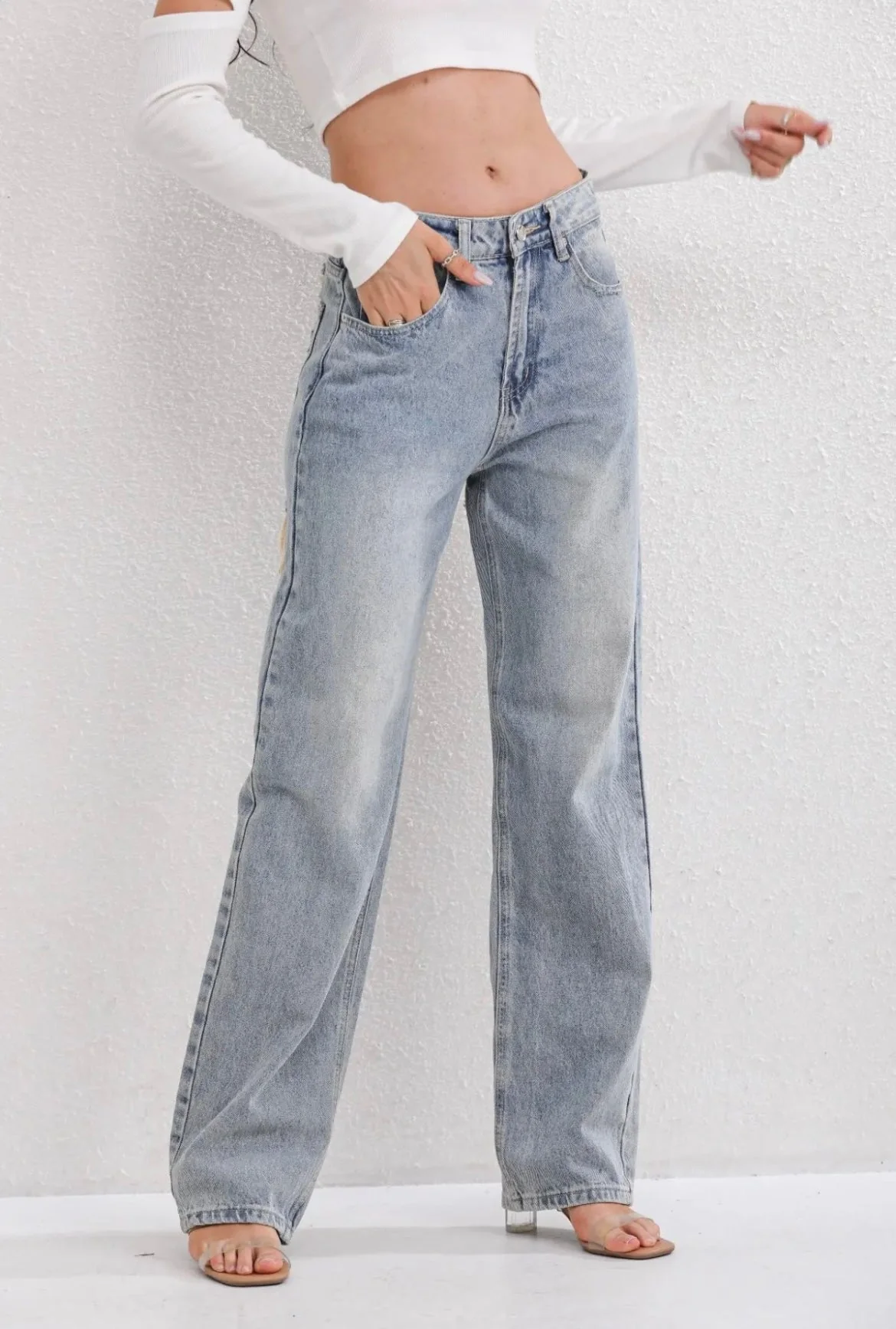 Jeune femme portant un jean large taille haute en denim bleu clair, associé à un haut blanc à manches longues, posant contre un mur texturé.