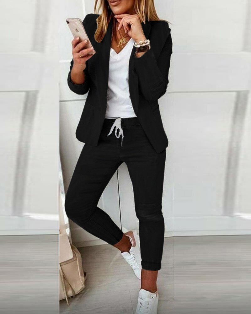 Femme en tenue décontractée chic, portant un blazer noir, t-shirt blanc, pantalon de jogging noir et baskets blanches, utilisant un smartphone.