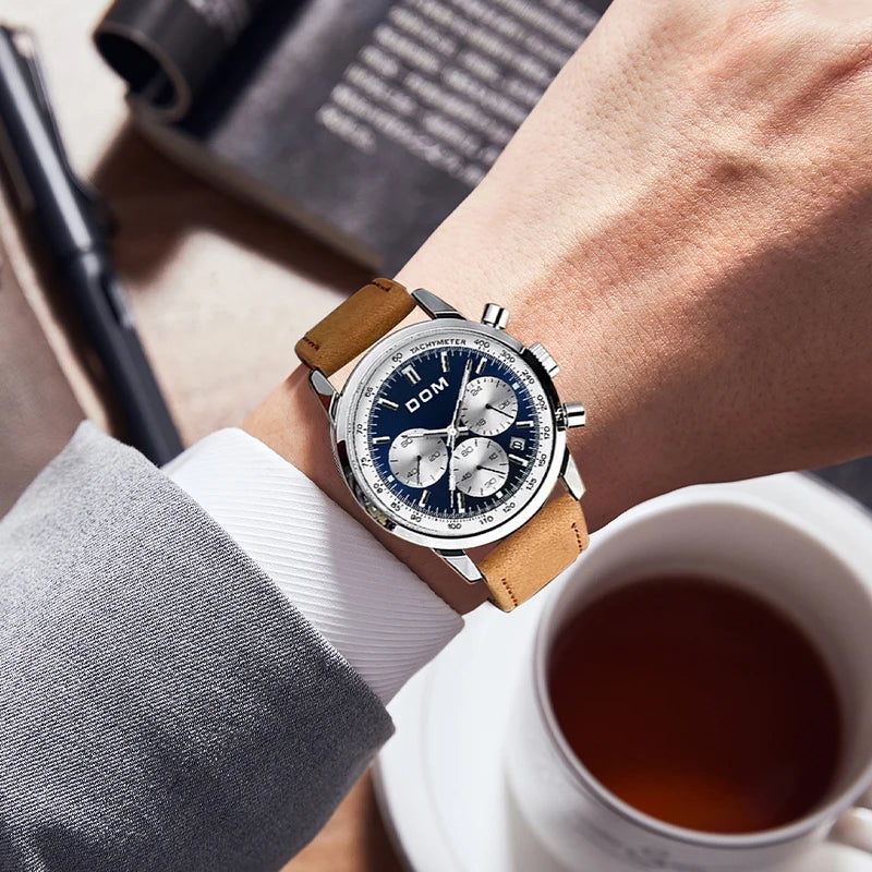 Montre-bracelet chronographe homme avec cadran bleu et bracelet en cuir marron, posée sur un poignet, à côté d'une tasse de thé et d'un livre.