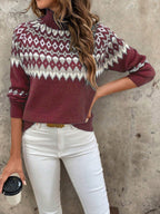 Femme portant un pull en laine bordeaux à motifs géométriques blancs, tenant une tasse de café, posant contre un mur gris. Mode automne-hiver tendance.