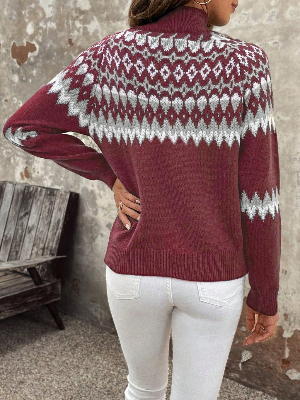 Femme portant un pull rouge à motifs géométriques blancs, manches longues, col montant, debout devant un mur texturé, style automne-hiver.
