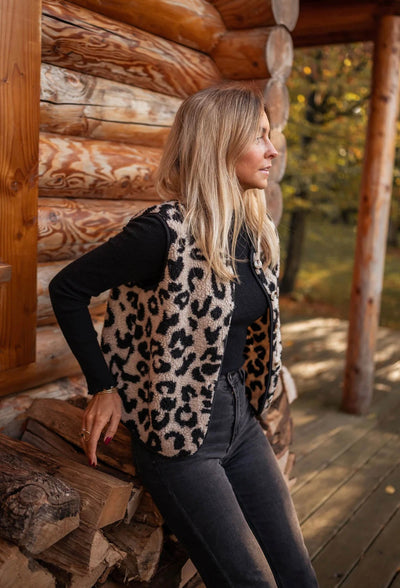 Femme blonde en gilet léopard, appuyée contre un mur en bois rond, regardant au loin. Mode automne, style décontracté, nature en arrière-plan.