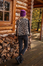 Femme en gilet léopard, bonnet violet, jeans noirs, bottes noires, devant une cabane en bois avec bûches, automne, forêt en arrière-plan.