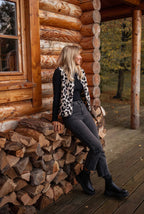 Femme blonde en gilet léopard, pull noir et jeans gris, assise sur un tas de bois devant une cabane en rondins, automne, style décontracté.