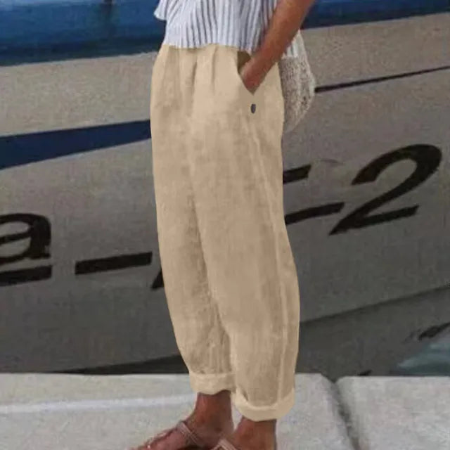 Personne portant un pantalon en lin beige, chemise rayée bleue et sandales marron, debout près d'un bateau. Mode estivale décontractée.