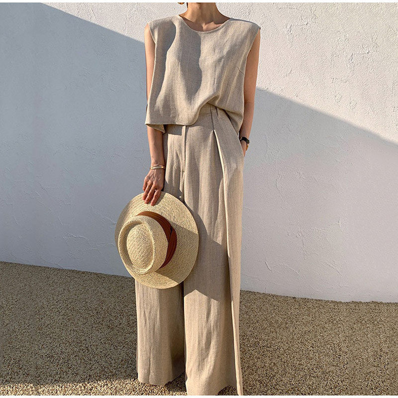 Femme en tenue beige élégante, tenant un chapeau de paille, posant contre un mur blanc. Mode estivale, style minimaliste, vêtements en lin.