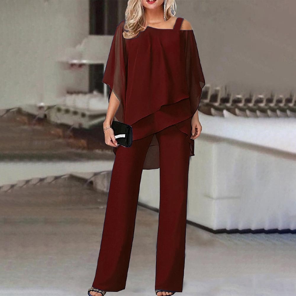 Femme en ensemble pantalon rouge bordeaux élégant, avec haut asymétrique et pochette noire, dans un intérieur moderne. Mode chic et tendance.