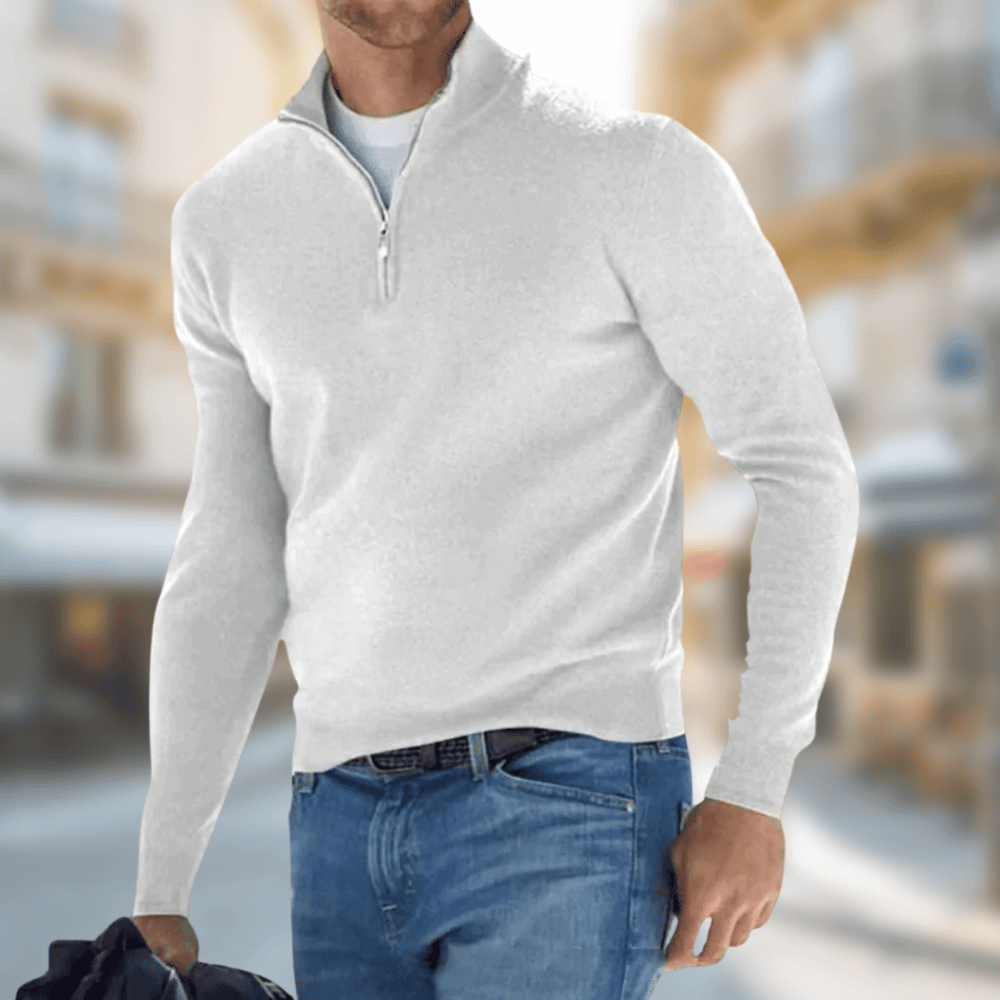 Homme portant un pull blanc à col zippé et un jean bleu, tenant une veste noire, dans une rue urbaine floue. Mode masculine élégante.