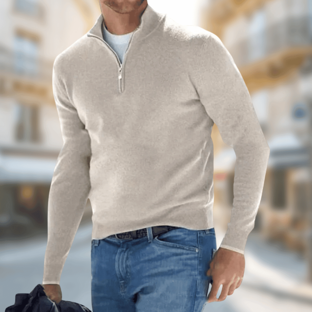 Homme portant un pull beige à col zippé, tenant un sac noir, dans une rue floue. Mode masculine, style décontracté, vêtements d'automne.