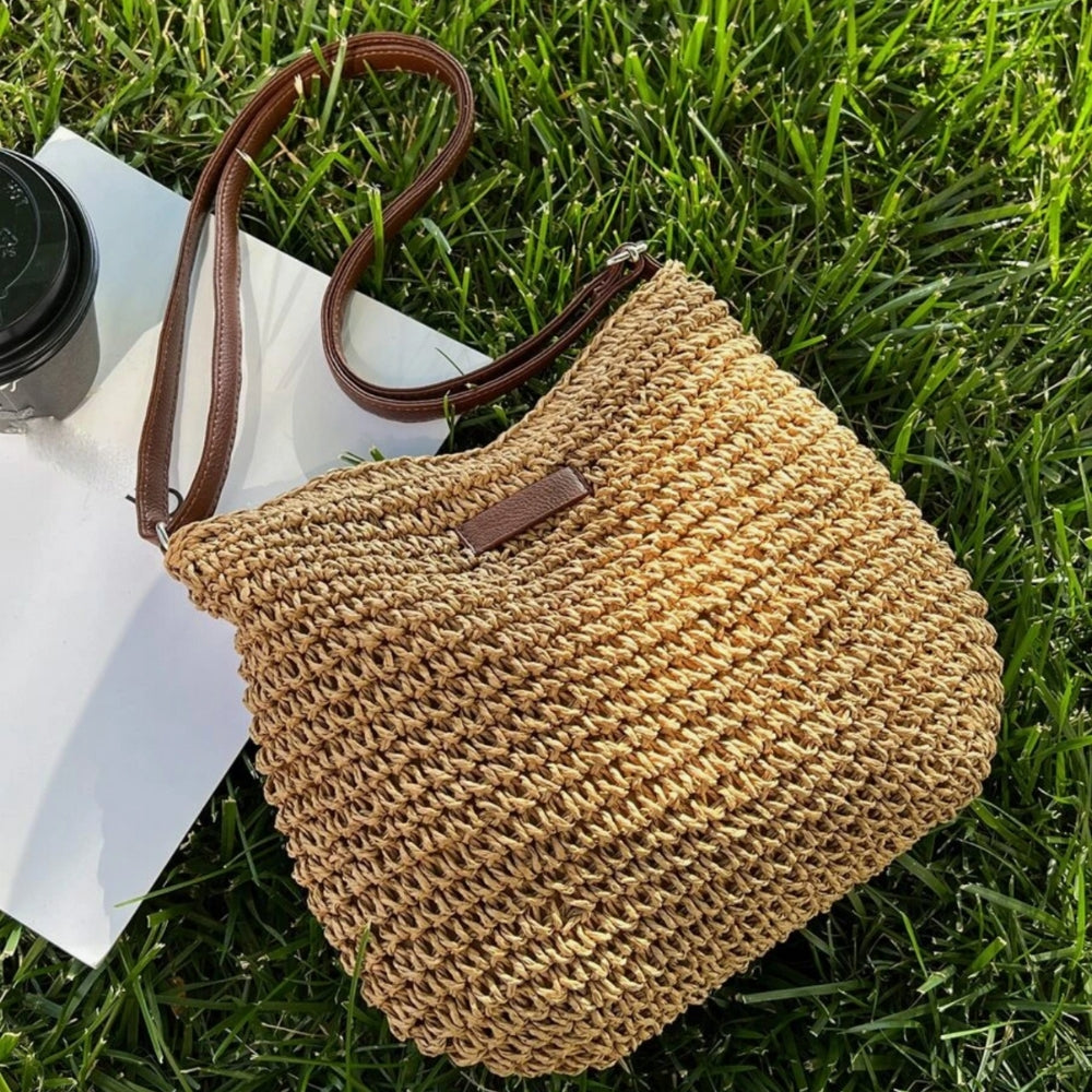 Sac en paille tressée sur herbe verte, avec bandoulière en cuir marron, à côté d'un gobelet noir. Accessoire estival tendance et naturel.