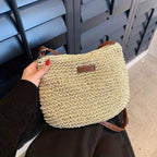 Sac à main en paille tressée beige avec bandoulière marron, tenu par une main avec vernis rouge, devant des volets noirs. Accessoire mode tendance.