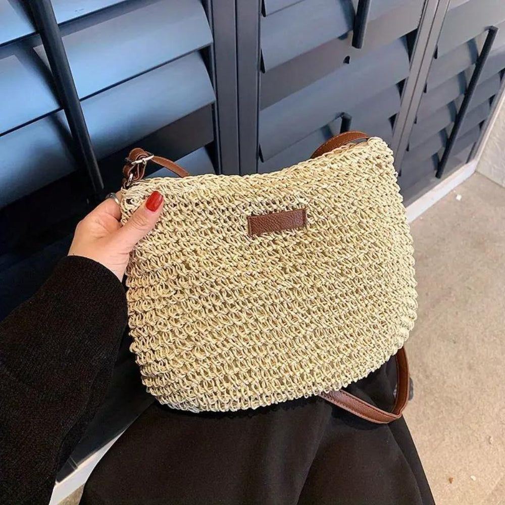 Sac à main en paille tressée beige avec bandoulière marron, tenu par une main avec vernis rouge, devant des volets noirs. Accessoire mode tendance.