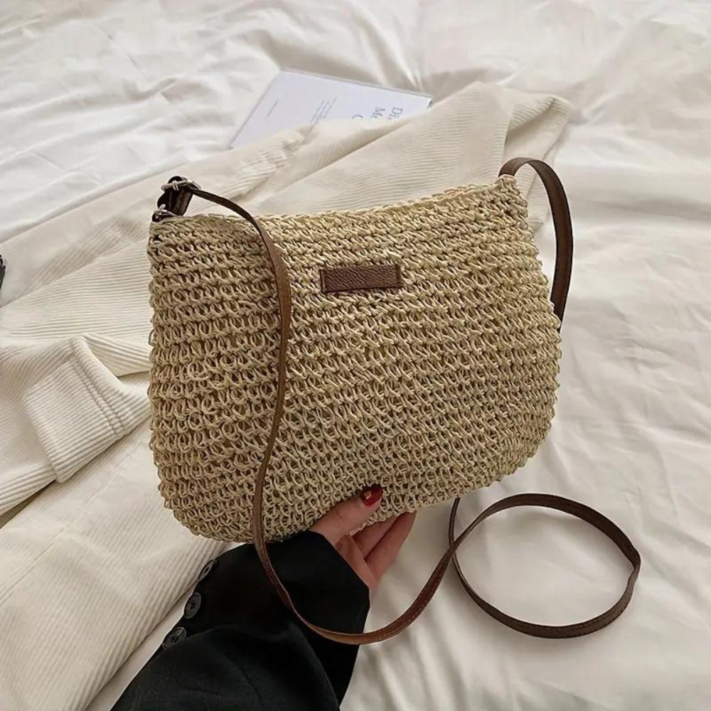 Sac à main en paille tressée beige avec bandoulière marron, posé sur un lit blanc. Accessoire mode tendance, idéal pour l'été et les sorties décontractées.