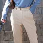 Personne portant un pull bleu et un pantalon beige élégant, tenant un sac à main marron. Mode automne, style décontracté chic, mur en pierre grise.