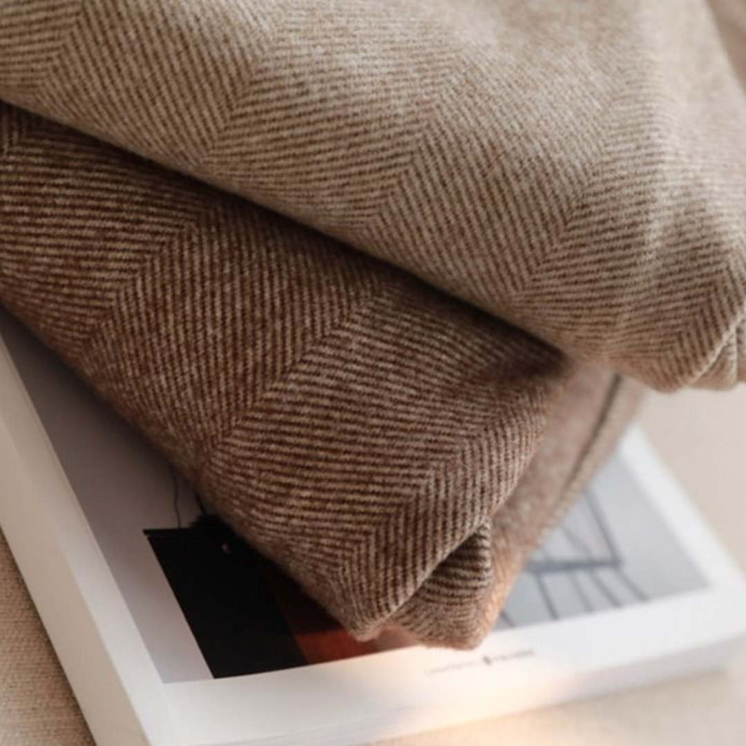 Deux couvertures en laine beige à motifs chevrons, empilées sur un livre, créant une ambiance chaleureuse et élégante pour la décoration intérieure.