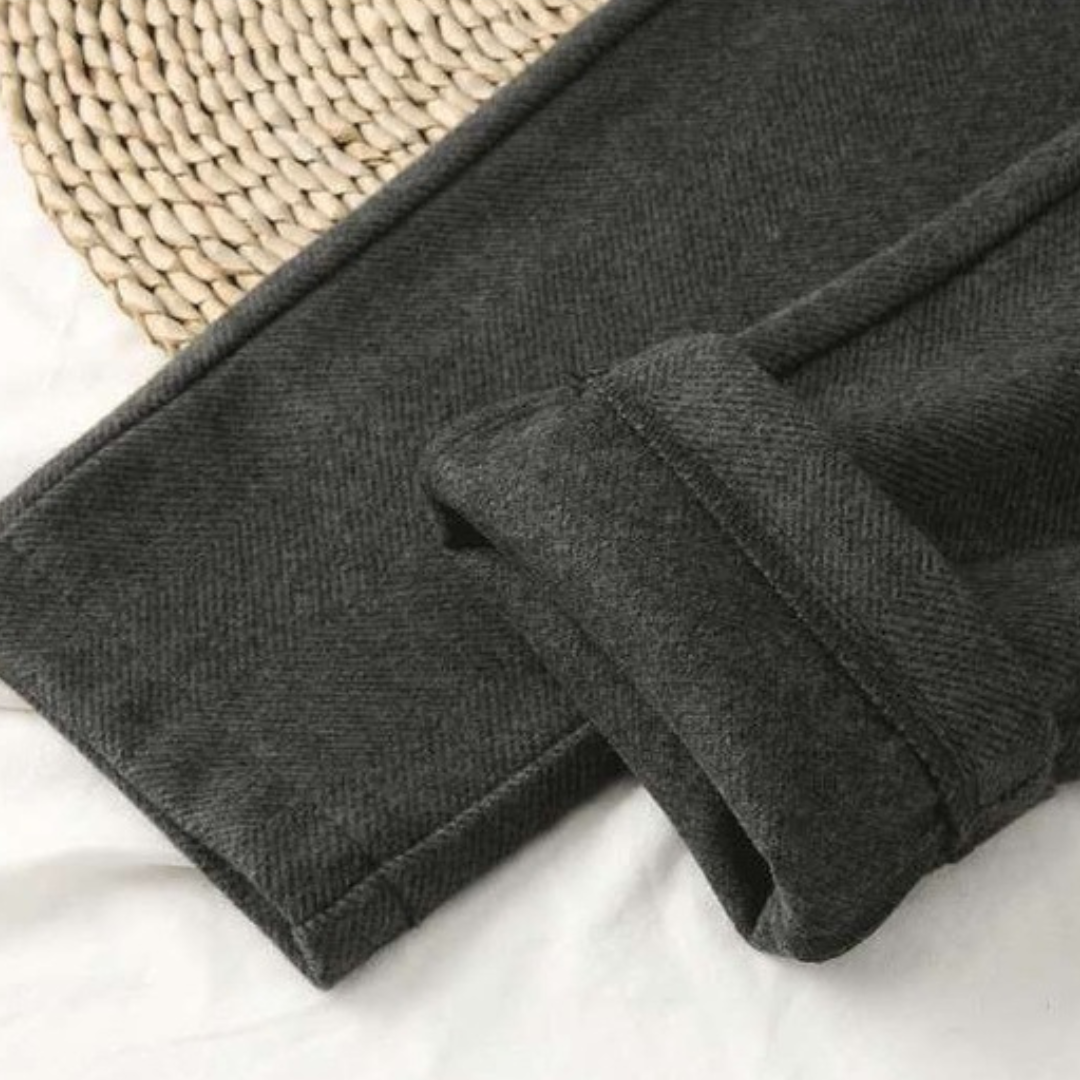 Pantalon en laine gris foncé, texture chevron, posé sur un tapis en osier. Mode hiver, vêtement chaud, style élégant et confortable.