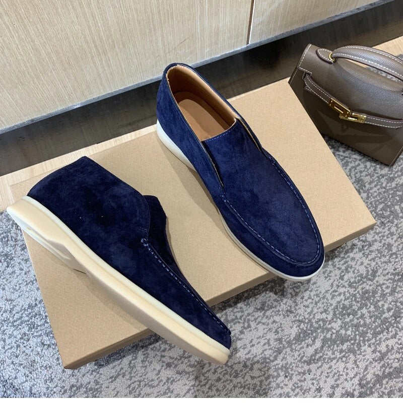 Mocassins en daim bleu marine pour homme sur boîte en carton, posés sur tapis gris, avec sac à main taupe en arrière-plan. Chaussures élégantes et modernes.